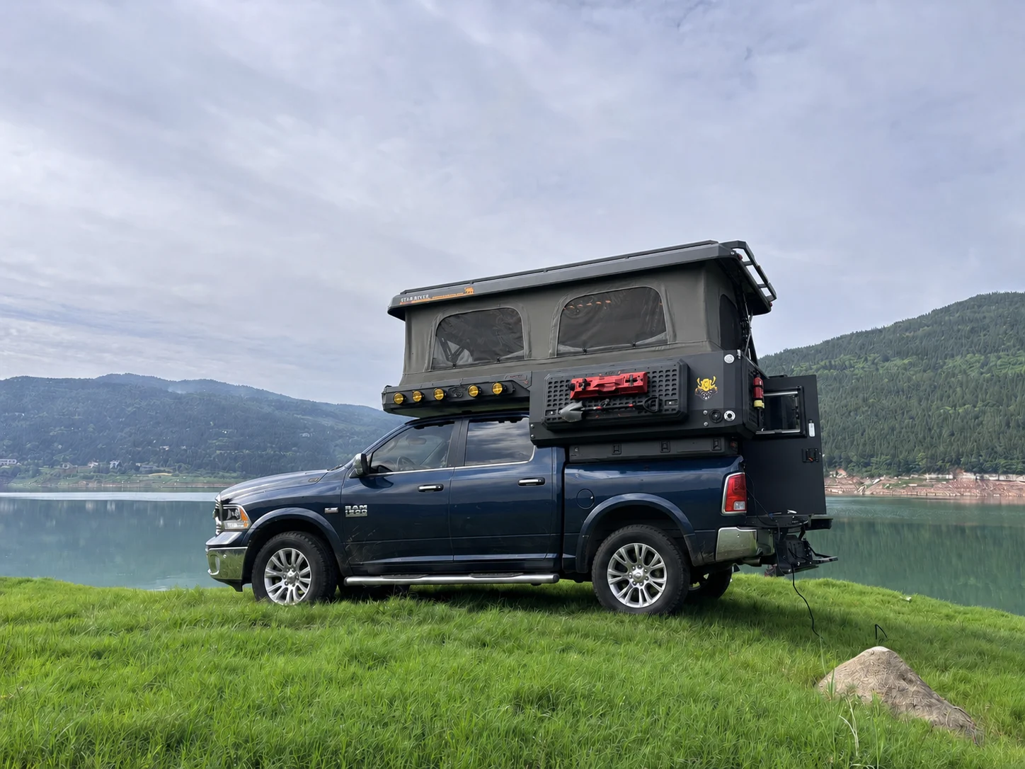 Blue RAM with a SUNHOMEE slide-in camper roof raised beside a mountain lake