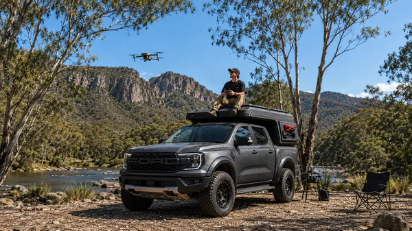 Grey pickup with a slide-in camper below sandstone cliffs, aerial view